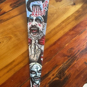 May include: A hand-painted metal hand saw featuring detailed portraits of two characters, one with a beard and one with a cross on the forehead. The blade includes the text "RABBIT" and "RUN" in red and white, with a hand gesturing a middle finger.