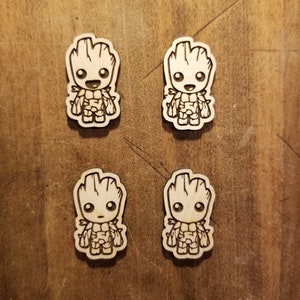 May include: Four wooden cutouts of a cartoon character, Groot from Guardians of the Galaxy, with a simple line drawing design. The cutouts are on a wood background.