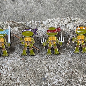 May include: Four Teenage Mutant Ninja Turtles enamel pins. Each pin features a different turtle with their signature weapon: Leonardo with katanas, Donatello with a bo staff, Raphael with sai, and Michelangelo with nunchakus.