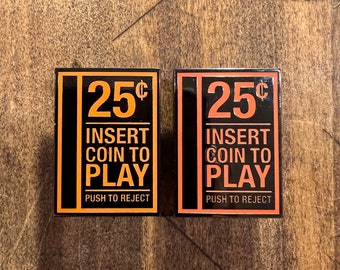 May include: Two black metal pinback buttons with orange text. The buttons read "25¢ Insert Coin to Play Push to Reject".