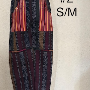 May include: Patchwork pants in shades of burgundy, navy, and brown with striped and patterned panels. The pants feature two pockets with vertical stripes in red, yellow, and orange. The text "#2 S/M" is visible on the right side.