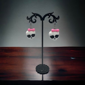 May include: A pair of white skull earrings with pink bows hanging from a black earring stand. The earrings are made of polymer clay.