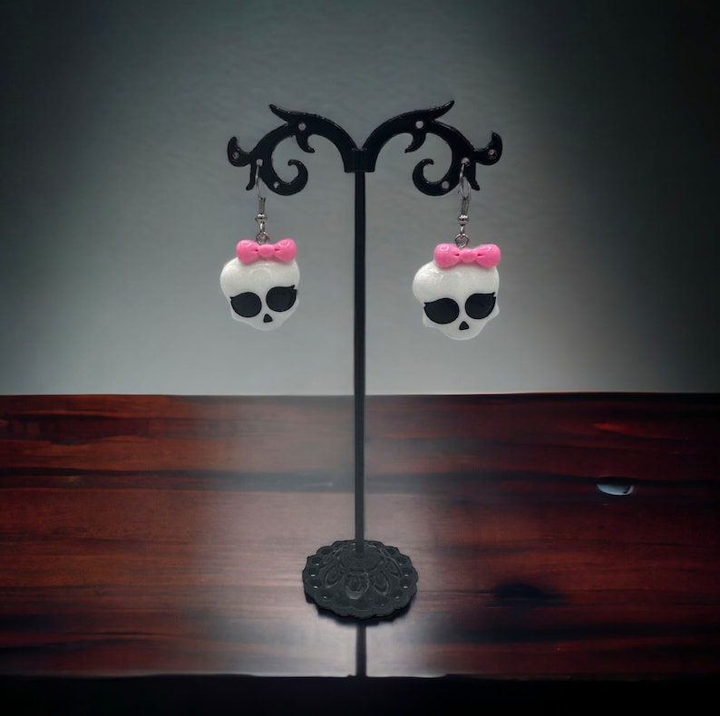 May include: A pair of white skull earrings with pink bows hanging from a black earring stand. The earrings are made of polymer clay.