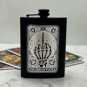 May include: Black hip flask featuring a silver graphic of a skeleton hand giving the middle finger, framed by roses and the text "THE ZERO OF FUCKS". The flask has a black screw-on cap.