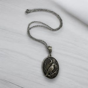 Puede incluir: Un collar con colgante ovalado de tono plateado que presenta un dise&ntilde;o en relieve de un cuervo posado sobre una rama bajo una luna creciente. El colgante cuelga de una delicada cadena plateada con una anilla texturizada.