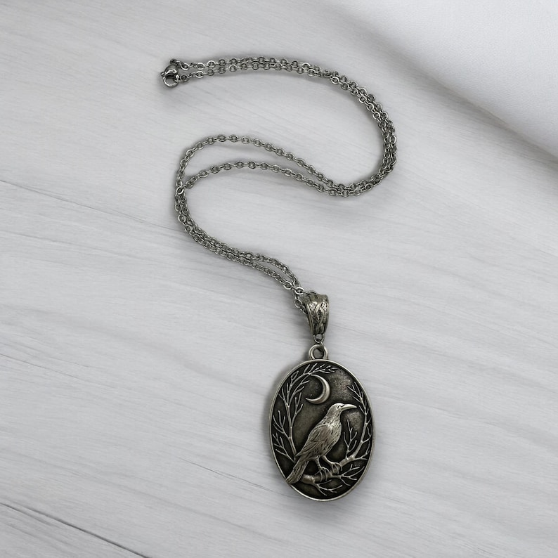 Puede incluir: Un collar con colgante ovalado de tono plateado que presenta un dise&ntilde;o en relieve de un cuervo posado sobre una rama bajo una luna creciente. El colgante cuelga de una delicada cadena plateada con una anilla texturizada.