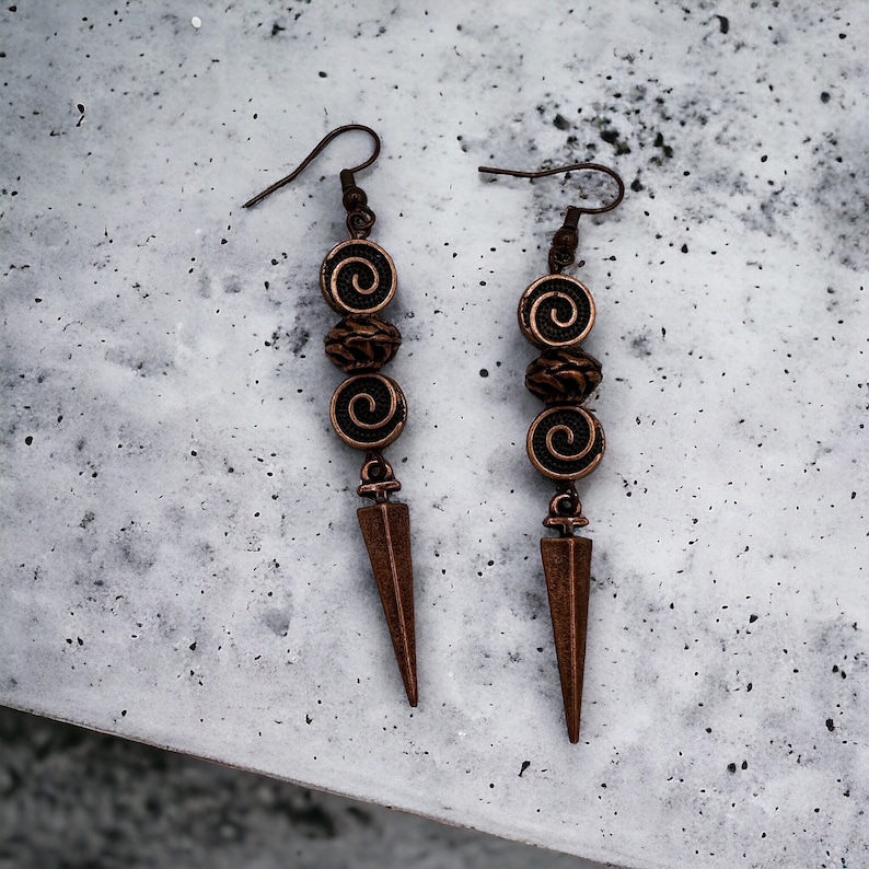 May include: A pair of long, copper-colored earrings with a pointed tip and three decorative beads. Each bead has a spiral design.