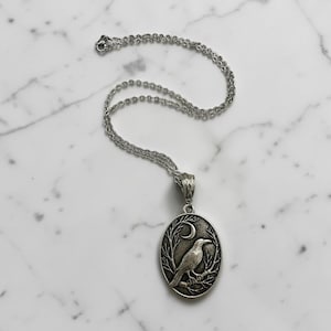 Puede incluir: Un collar con colgante ovalado de tono plateado que presenta un relieve detallado de un cuervo posado en una rama bajo una luna creciente. El colgante cuelga de una delicada cadena plateada con una anilla texturizada.
