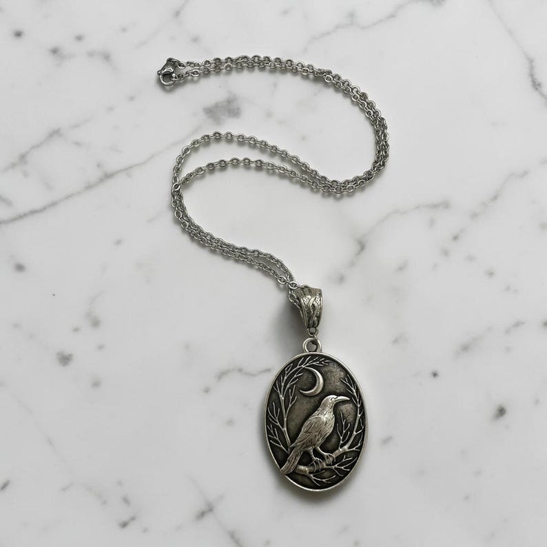 Puede incluir: Un collar con colgante ovalado de tono plateado que presenta un relieve detallado de un cuervo posado en una rama bajo una luna creciente. El colgante cuelga de una delicada cadena plateada con una anilla texturizada.