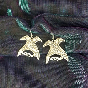 May include: A pair of silver earrings featuring a detailed design of two birds with outstretched wings. The earrings are on a black and green iridescent fabric background.