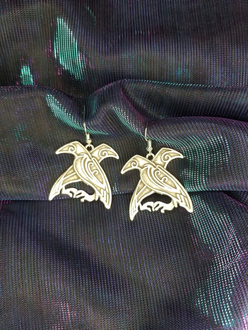 May include: A pair of silver earrings featuring a detailed design of two birds with outstretched wings. The earrings are on a black and green iridescent fabric background.