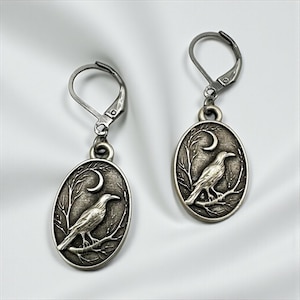May include: A pair of silver-toned oval drop earrings featuring a raised relief design of a raven perched on a bare tree branch under a crescent moon. The earrings are finished with secure lever-back closures, showcasing an antique metal texture.