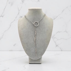 May include: A silver-toned lariat necklace featuring a central circular ring connector. Three delicate chains of varying lengths descend from the ring, each ending in a slender, pointed metal spike pendant with a textured, etched surface finish.
