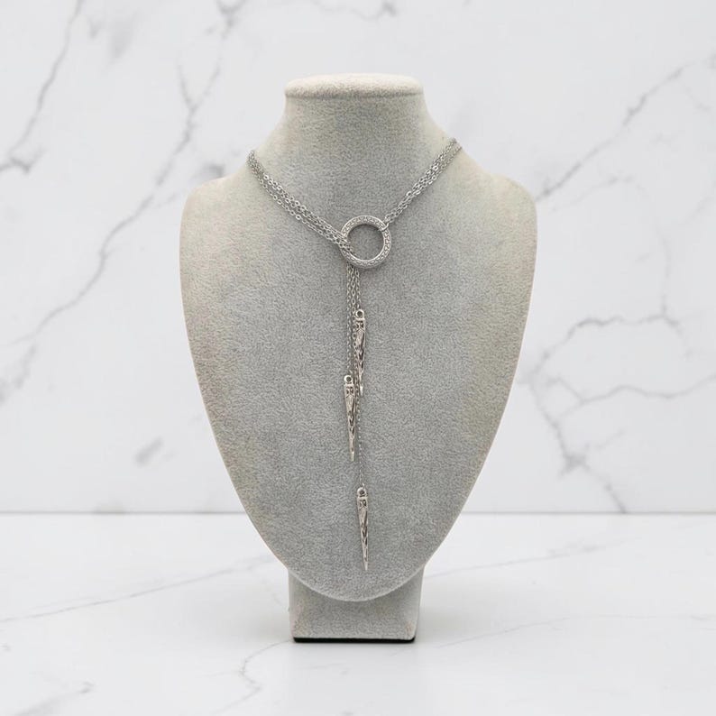 May include: A silver-toned lariat necklace featuring a central circular ring connector. Three delicate chains of varying lengths descend from the ring, each ending in a slender, pointed metal spike pendant with a textured, etched surface finish.