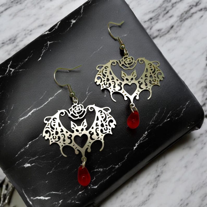 May include: A pair of silver earrings shaped like bats with intricate cutouts and red glass drops. The bats have a heart-shaped design in the center and a rose detail on the top.