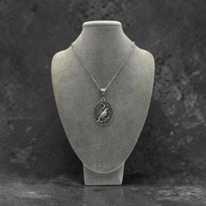 Puede incluir: Un collar de tono plateado con un colgante ovalado que presenta un relieve detallado de un p&aacute;jaro posado en una rama bajo una luna creciente. El colgante cuelga de una delicada cadena plateada, mostrando un acabado met&aacute;lico pulido y grabado.