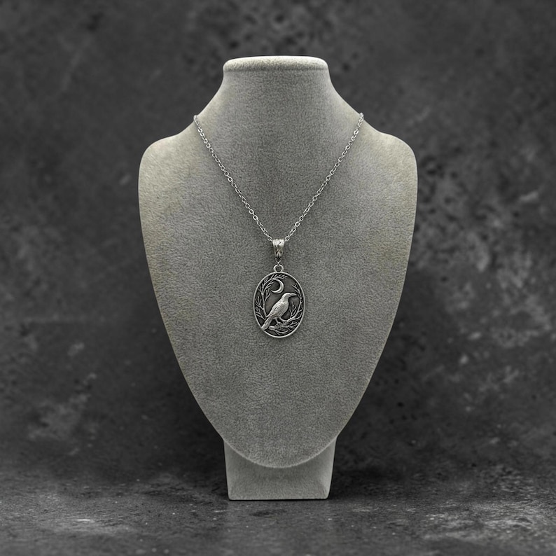 Puede incluir: Un collar de tono plateado con un colgante ovalado que presenta un relieve detallado de un p&aacute;jaro posado en una rama bajo una luna creciente. El colgante cuelga de una delicada cadena plateada, mostrando un acabado met&aacute;lico pulido y grabado.
