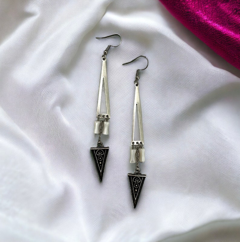 Puede incluir: Un par de pendientes de plata con un dise&ntilde;o triangular. Los pendientes tienen un patr&oacute;n geom&eacute;trico negro y cuentas blancas.