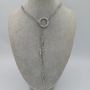 May include: A silver-toned lariat necklace featuring a circular pendant with a Greek key pattern. The necklace has multiple delicate chains that drape downward, each ending in a slender, pointed metal charm with intricate engraved detailing.