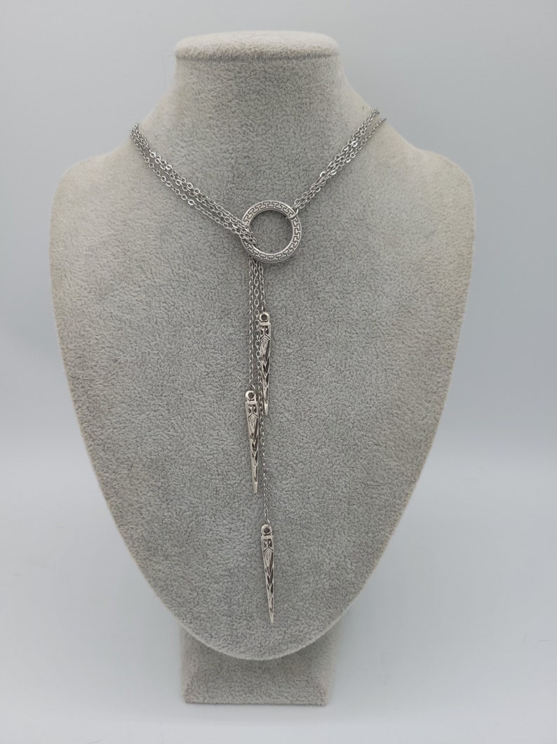 May include: A silver-toned lariat necklace featuring a circular pendant with a Greek key pattern. The necklace has multiple delicate chains that drape downward, each ending in a slender, pointed metal charm with intricate engraved detailing.
