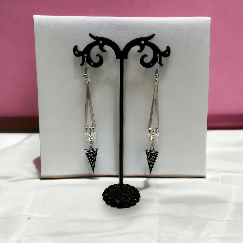 Puede incluir: Un par de pendientes colgantes de plata con colgantes triangulares negros y cuentas blancas que cuelgan de una cadena de plata. Los pendientes se muestran en un soporte negro.