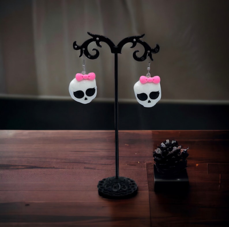 May include: A pair of white skull earrings with pink bows hanging on a black earring stand. The earrings are on a wooden surface.