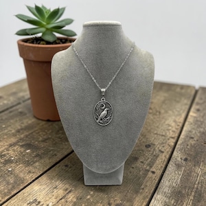 Puede incluir: Un collar de tono plateado con un colgante ovalado que presenta un dise&ntilde;o en relieve de un p&aacute;jaro posado en una rama bajo una luna creciente. El colgante cuelga de una delicada cadena plateada, mostrando un trabajo de metal detallado.