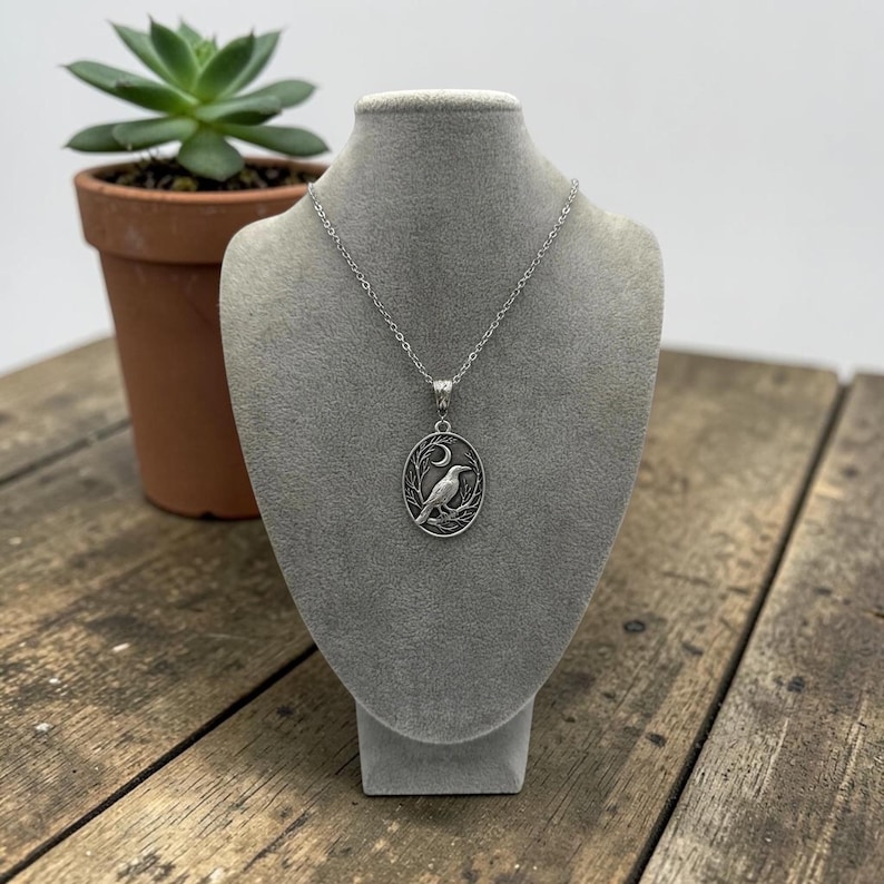Puede incluir: Un collar de tono plateado con un colgante ovalado que presenta un dise&ntilde;o en relieve de un p&aacute;jaro posado en una rama bajo una luna creciente. El colgante cuelga de una delicada cadena plateada, mostrando un trabajo de metal detallado.