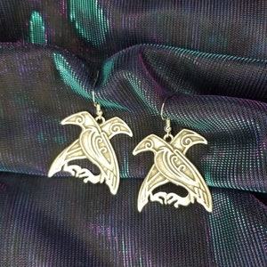 May include: A pair of silver earrings featuring a detailed design of a bird with intricate patterns. The earrings are displayed on a iridescent fabric background.
