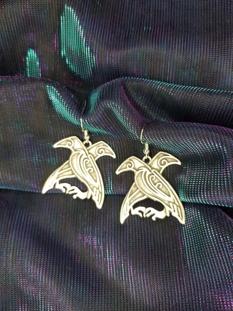 May include: A pair of silver earrings featuring a detailed design of a bird with intricate patterns. The earrings are displayed on a iridescent fabric background.