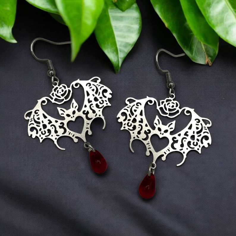 May include: Silver earrings shaped like bats with a rose detail and dangling red teardrop crystals.