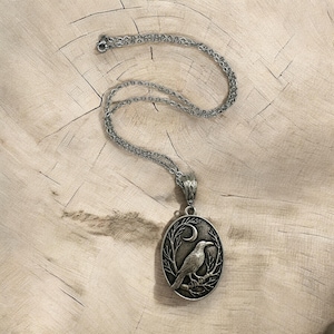Puede incluir: Un collar con colgante ovalado de tono plateado que presenta un dise&ntilde;o en relieve de un cuervo posado sobre una rama bajo una luna creciente. El colgante cuelga de una delicada cadena plateada a juego con una estructura de eslabones.