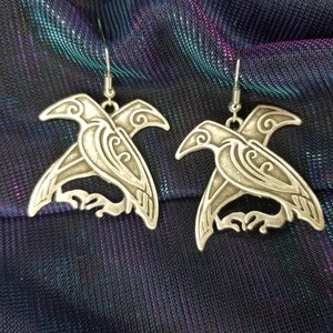 May include: A pair of silver earrings featuring a detailed design of a bird with intricate patterns. The earrings are hanging from silver hooks.