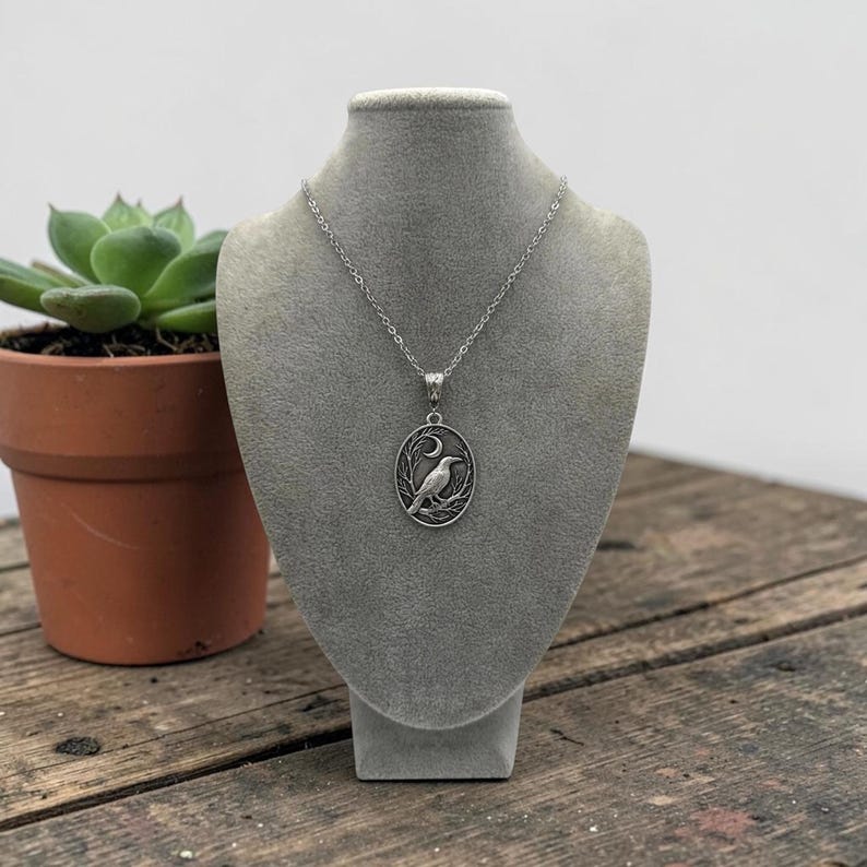 Puede incluir: Un collar con colgante ovalado de tono plateado que presenta un dise&ntilde;o en relieve de un p&aacute;jaro posado en una rama bajo una luna creciente. El colgante cuelga de una fina cadena plateada, exhibido sobre un busto de terciopelo gris.