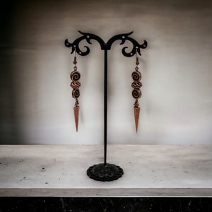 May include: A pair of long, gold-toned earrings with a pointed tip and three spiral-shaped beads hanging from each earring. The earrings are displayed on a black stand.