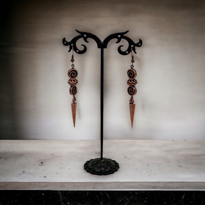 May include: A pair of long, gold-toned earrings with a pointed tip and three spiral-shaped beads hanging from each earring. The earrings are displayed on a black stand.