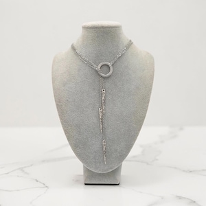 May include: A silver-toned lariat necklace featuring a central circular ring connector. Three delicate chains extend downward from the ring, each terminating in a slender, pointed metal spike charm. The necklace is displayed on a neutral bust.