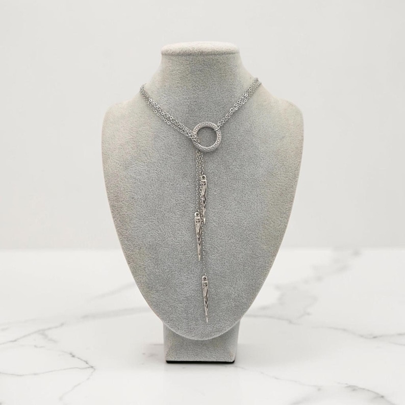 May include: A silver-toned lariat necklace featuring a central circular ring connector. Three delicate chains extend downward from the ring, each terminating in a slender, pointed metal spike charm. The necklace is displayed on a neutral bust.
