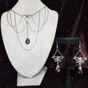 May include: A silver chain necklace with a dangling crystal pendant and matching earrings. The necklace has a delicate, intricate design and the crystal is a deep purple color.