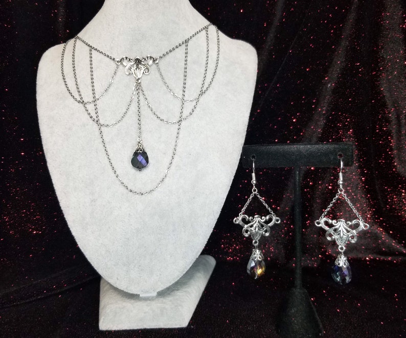 May include: A silver chain necklace with a dangling crystal pendant and matching earrings. The necklace has a delicate, intricate design and the crystal is a deep purple color.