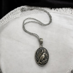 Puede incluir: Un collar con colgante ovalado de tono plateado que presenta un relieve detallado de un cuervo posado en una rama bajo una luna creciente. El colgante cuelga de una delicada cadena plateada a juego con un pasador texturizado y adornado.