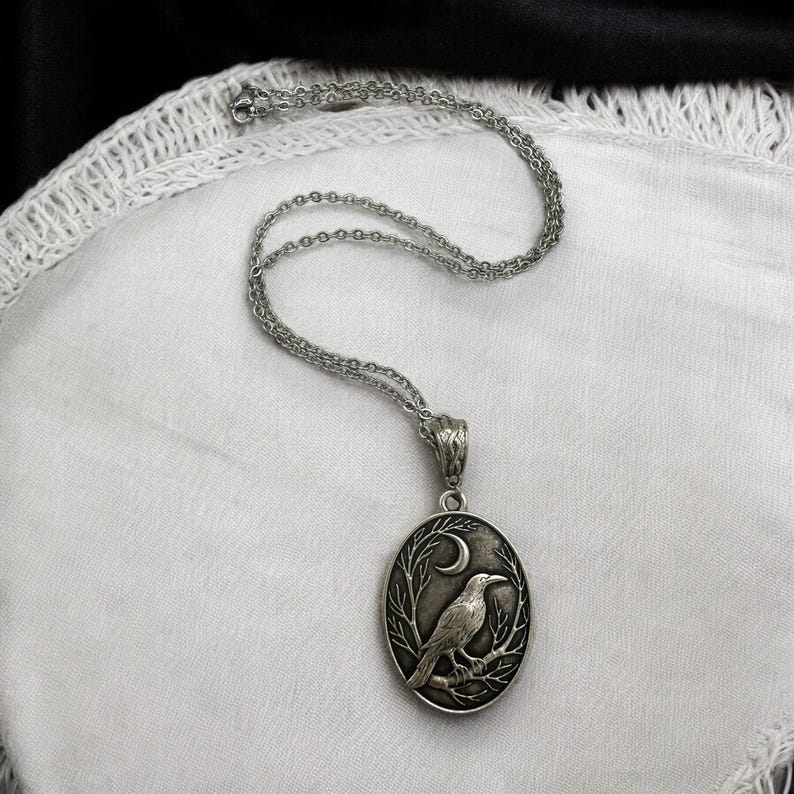 Puede incluir: Un collar con colgante ovalado de tono plateado que presenta un relieve detallado de un cuervo posado en una rama bajo una luna creciente. El colgante cuelga de una delicada cadena plateada a juego con un pasador texturizado y adornado.