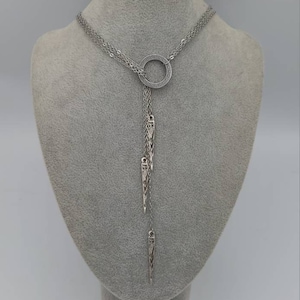 May include: A silver-toned lariat necklace featuring a central circular ring encrusted with small clear stones. Three delicate chains descend from the ring, each ending in a slender, pointed metal pendant with intricate engraved detailing.