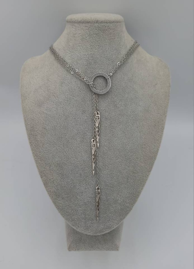 May include: A silver-toned lariat necklace featuring a central circular ring encrusted with small clear stones. Three delicate chains descend from the ring, each ending in a slender, pointed metal pendant with intricate engraved detailing.