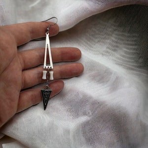 Puede incluir: Un pendiente de plata en forma de tri&aacute;ngulo con un dije blanco similar al hueso y un dije triangular m&aacute;s peque&ntilde;o con detalles negros.