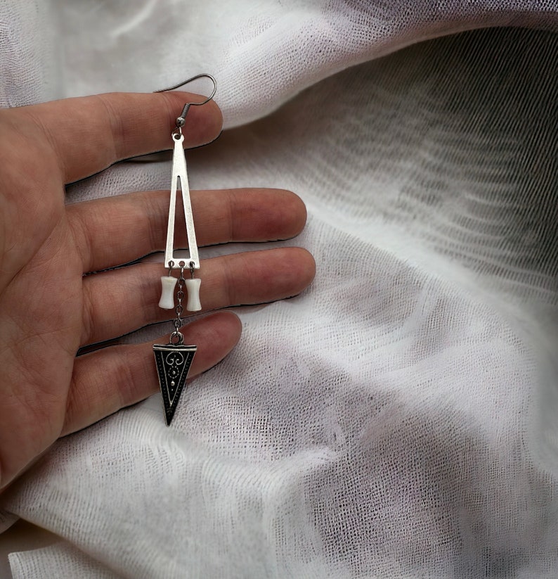 Puede incluir: Un pendiente de plata en forma de tri&aacute;ngulo con un dije blanco similar al hueso y un dije triangular m&aacute;s peque&ntilde;o con detalles negros.