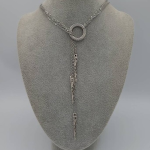 May include: A silver-toned lariat necklace featuring a circular pendant with a Greek key pattern. Three delicate chains hang from the ring, each ending in a slender, pointed metal charm with intricate engraved detailing along the surface.