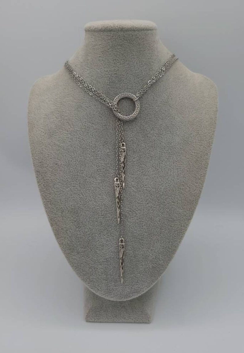 May include: A silver-toned lariat necklace featuring a circular pendant with a Greek key pattern. Three delicate chains hang from the ring, each ending in a slender, pointed metal charm with intricate engraved detailing along the surface.