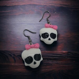 May include: A pair of white skull earrings with black eyes and pink bows. The earrings are made of polymer clay and have silver hooks.