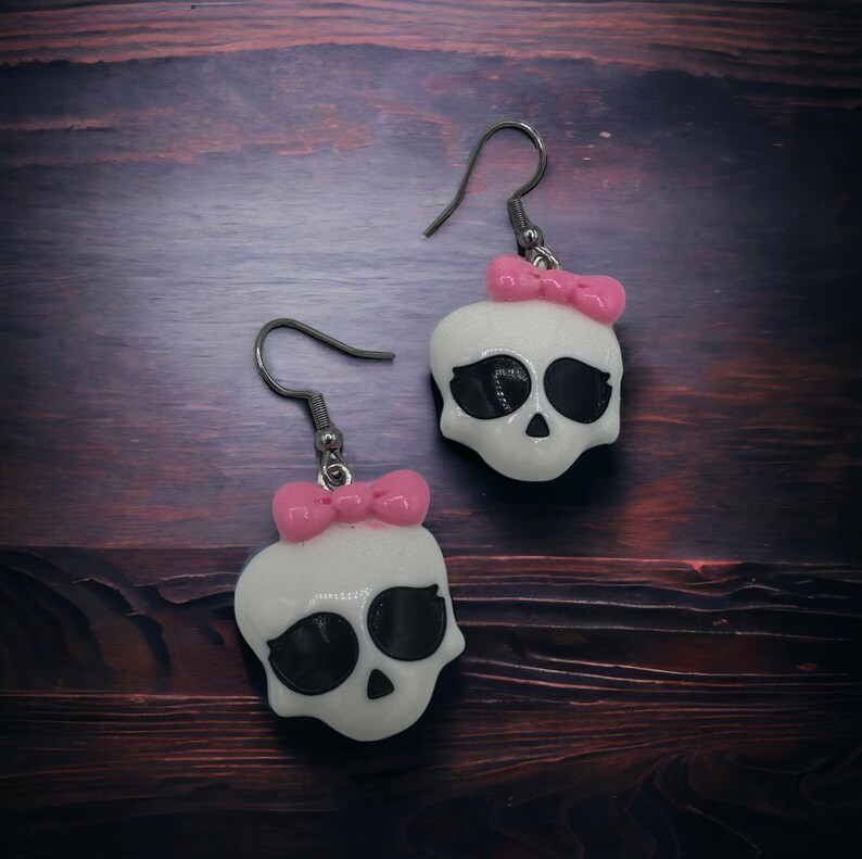 May include: A pair of white skull earrings with black eyes and pink bows. The earrings are made of polymer clay and have silver hooks.